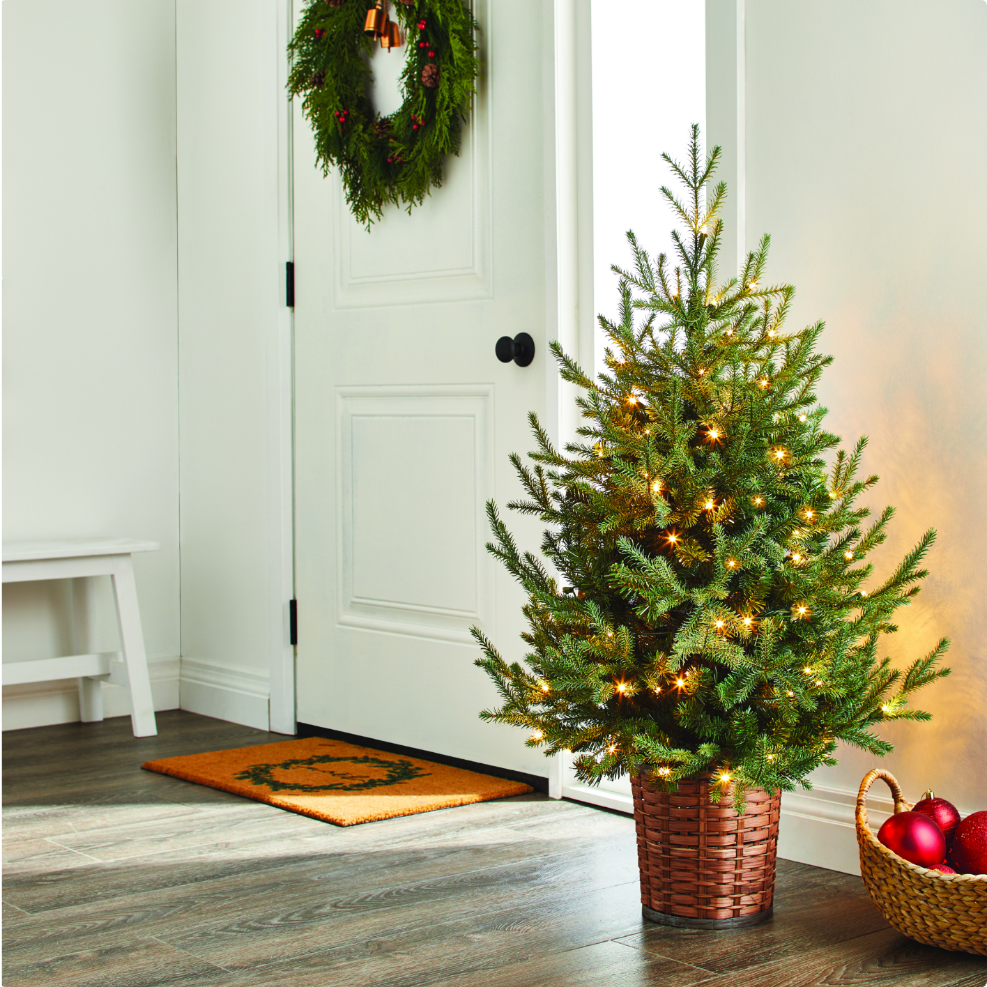 Une entrée avec un sapin de Noël illuminé, une couronne sur la porte, un paillasson et un panier rempli d'ornements rouges posé au sol.  