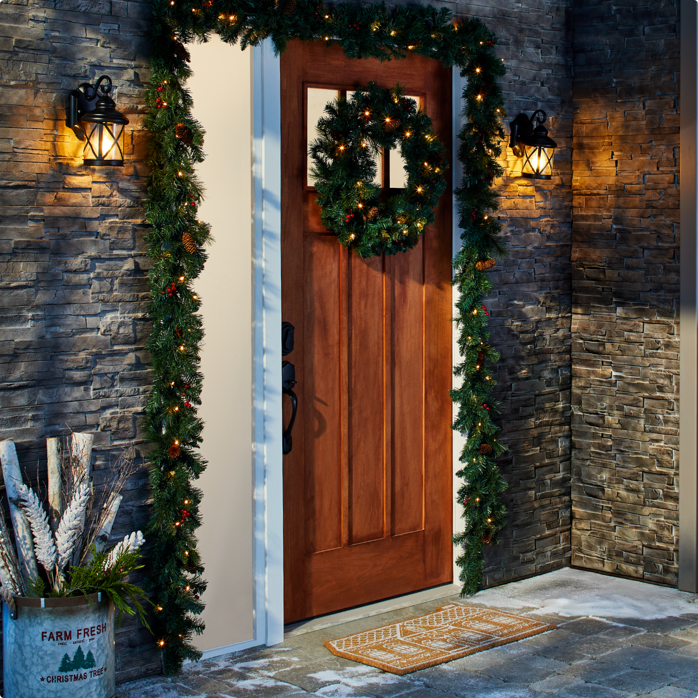 Une porte d'entrée ornée d'une couronne, de guirlandes lumineuses, d'un paillasson et d'un seau métallique rempli de bûches de bouleau.
