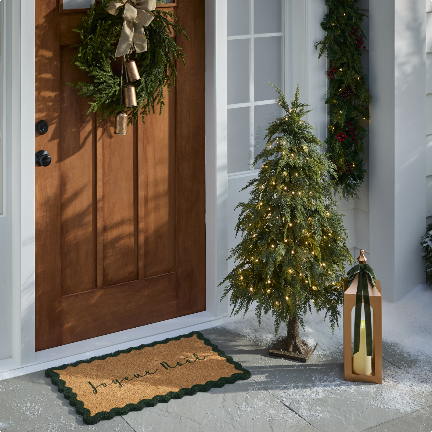 Une porte d'entrée ornée d'une couronne, d'un petit sapin illuminé, d'un paillasson sur le thème de Noël et d'une lanterne avec des bougies sur le porche.