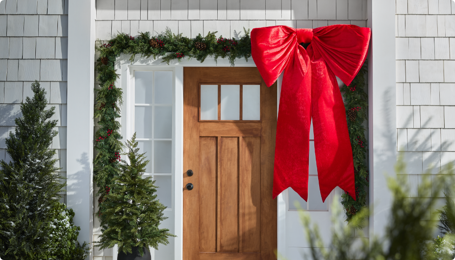 Une porte d'entrée ornée d'une grande boucle rouge, de guirlandes et de petits sapins de Noël de chaque côté. 
