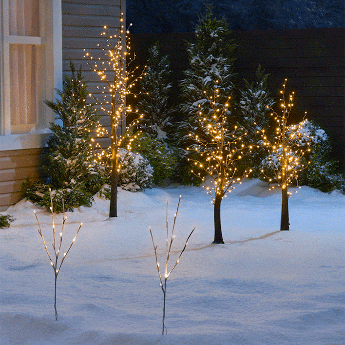 Snowy outdoor scene with small trees and bushes decorated with warm, glowing holiday lights.