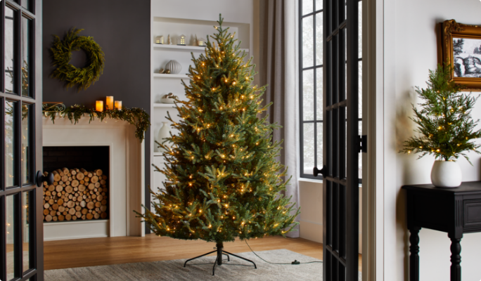 Lit Christmas tree in a living room with a fireplace, wreath, candles, and holiday decor on shelves and a table.