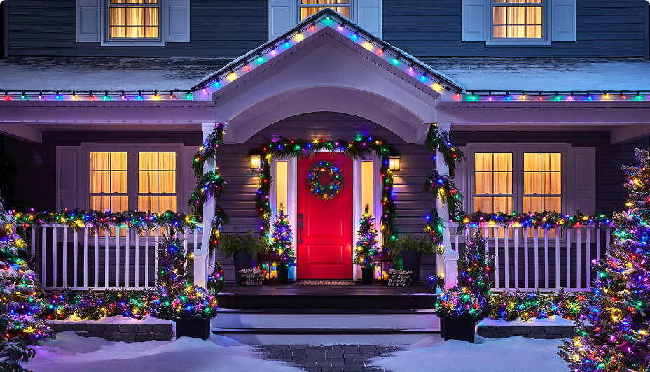 House decorated with colorful Christmas lights, wreath on the red door, and snow-covered ground.