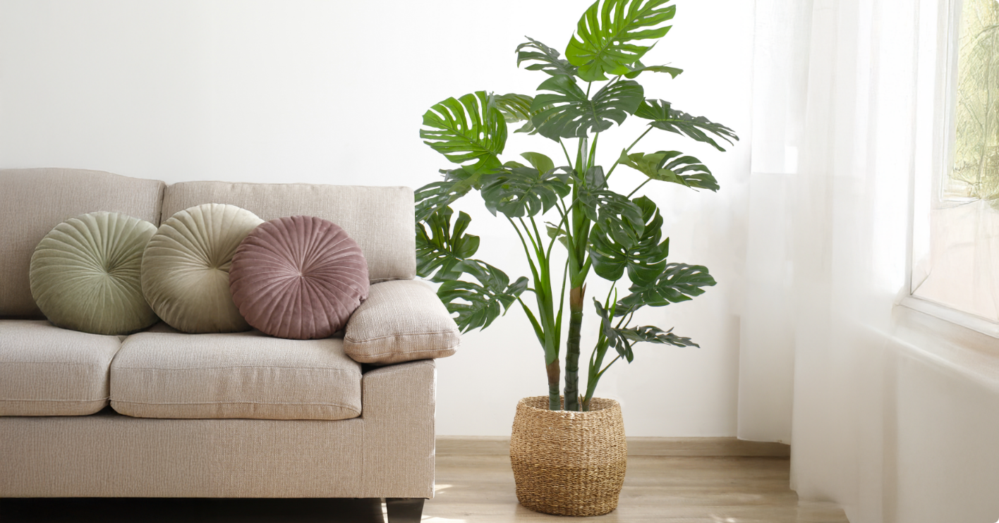 A large artificial potted plant next to a beige couch in a bright, minimalist room.