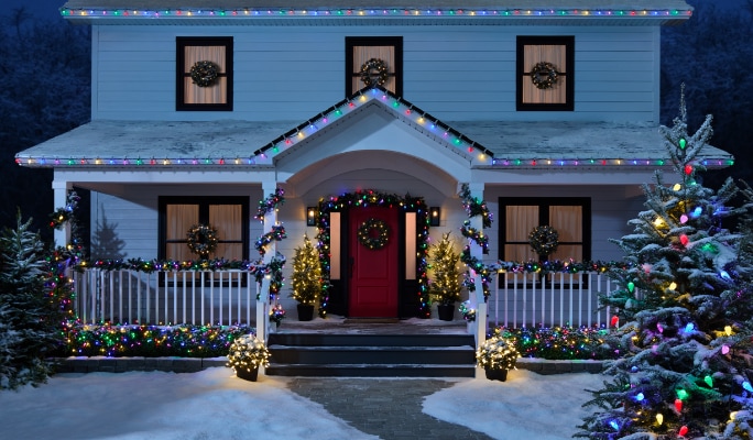 A house decorated with NOMA Advanced EverTough Christmas lights, wreaths on windows, a lit-up tree, and snow on the ground.