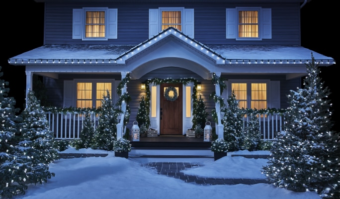 A house with warm lights in the windows, Christmas decorations, wreaths, garlands, lit-up trees, and snow on the ground.