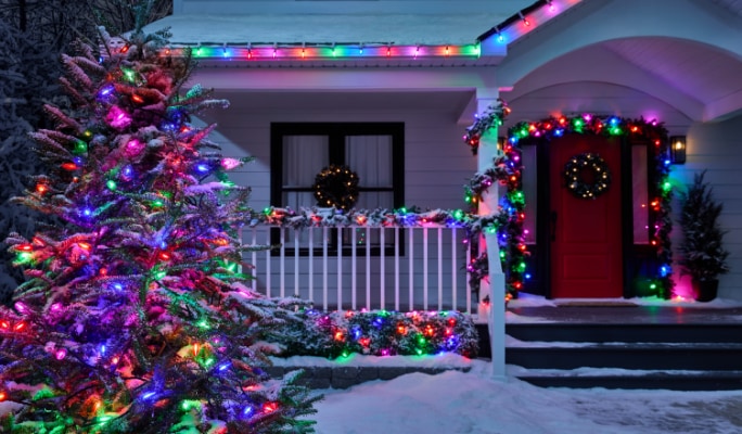 A house bathed in the purple light of Big & Bright Christmas lights, a lit-up tree, garlands, and wreaths, with snow on the ground.