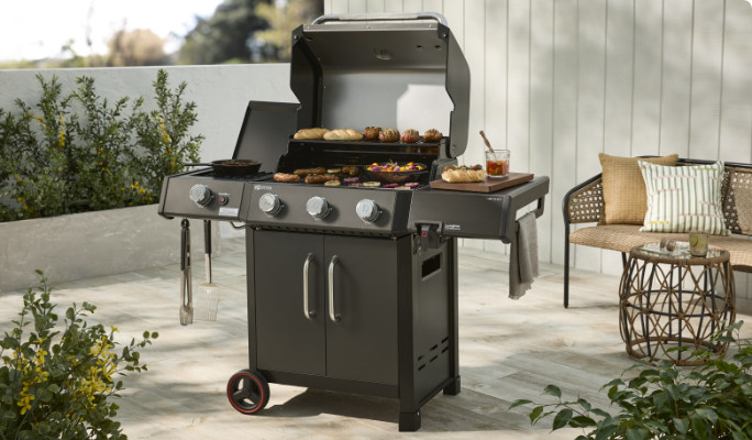 A Coleman Revolution ll 3-Burner BBQ cooking foods on a patio in front of a patio loveseat and coffee table.