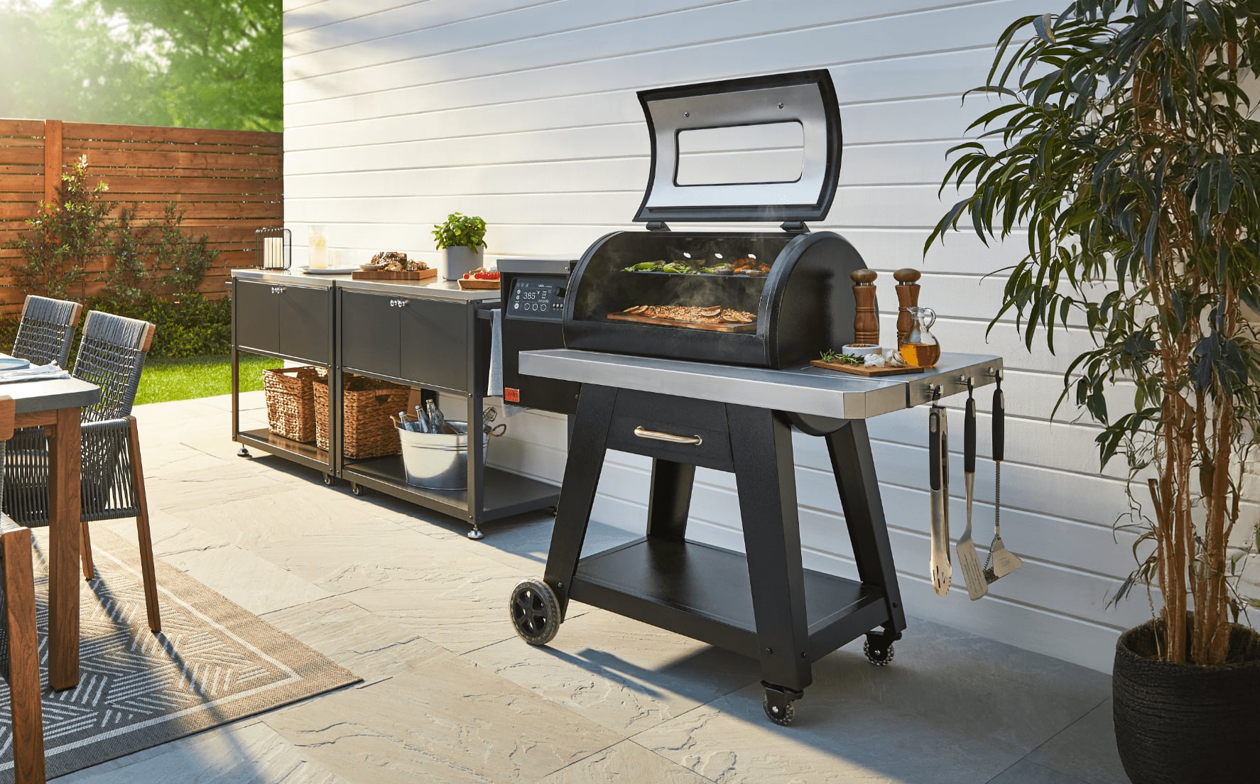 A Vida by PADERNO Wi-Fi & Bluetooth Connected Pellet Grill on a sunny patio next to two bar carts, plants and a patio dining table set.