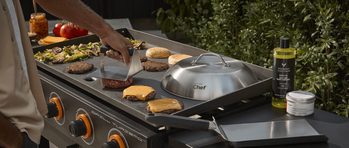 A person pressing a burger on a griddle top filled with cheeseburgers and vegetables.