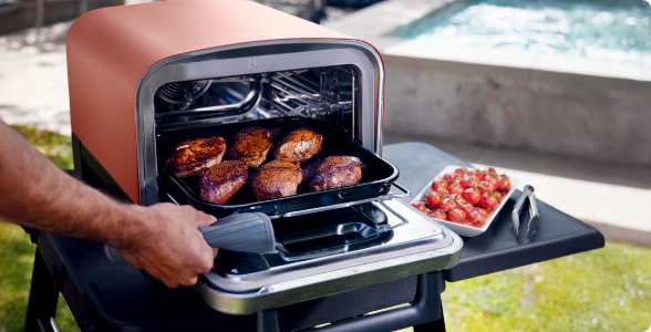 A Ninja Woodfire™ Outdoor Oven cooking food on a table, outside by a pool.
