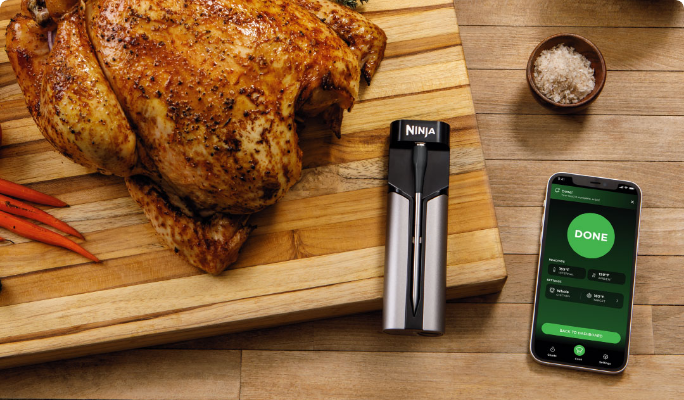 A Ninja ProChef Wireless Meat Thermometer next to a smart phone and a cooked turkey on a cutting board.