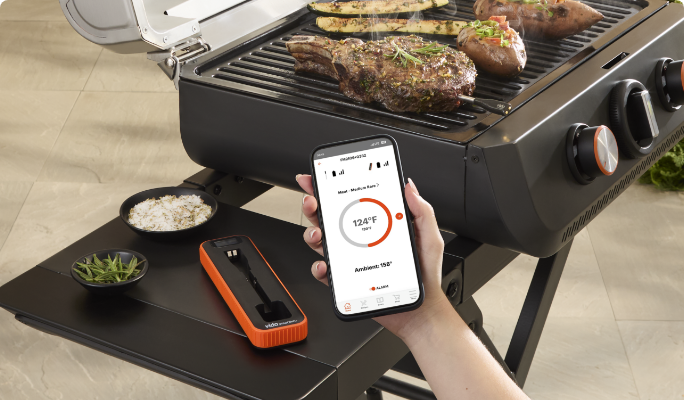 A Vida by PADERNO Smart Thermometer next to someone holding a smart phone by a grill cooking steaks.