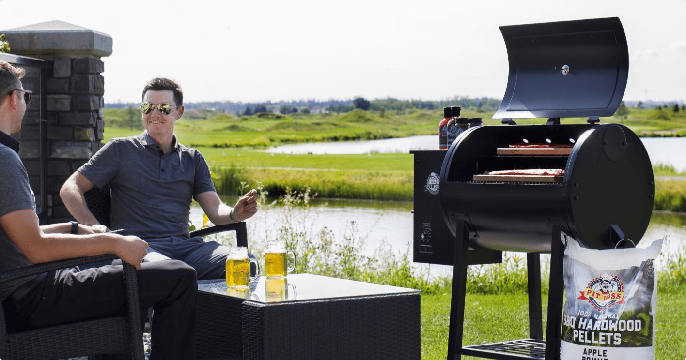 Two people sitting outside with drinks by a lake in front of a Pit Boss® 700FB1 Series Pellet Grill.