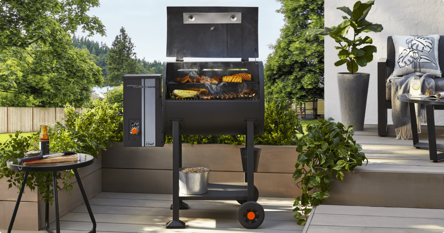 A MASTER Chef® Grill Turismo Pellet Grill & Smoker cooking various foods on a back patio surrounded by greenery.