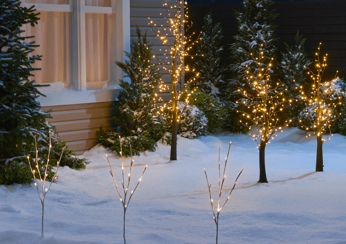 Divers arbres en fleurs scintillants CANVAS et lampes sur piquet bouleau scintillant CANVAS sont exposés dans une cour enneigée la nuit. 