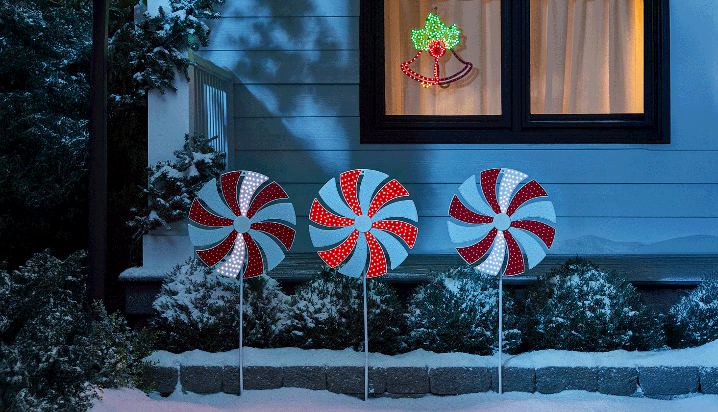 Cloches néon NOMA intérieures et extérieures et cannes en bonbon de Noël néon NOMA extérieures exposées dans une cour avant enneigée la nuit.