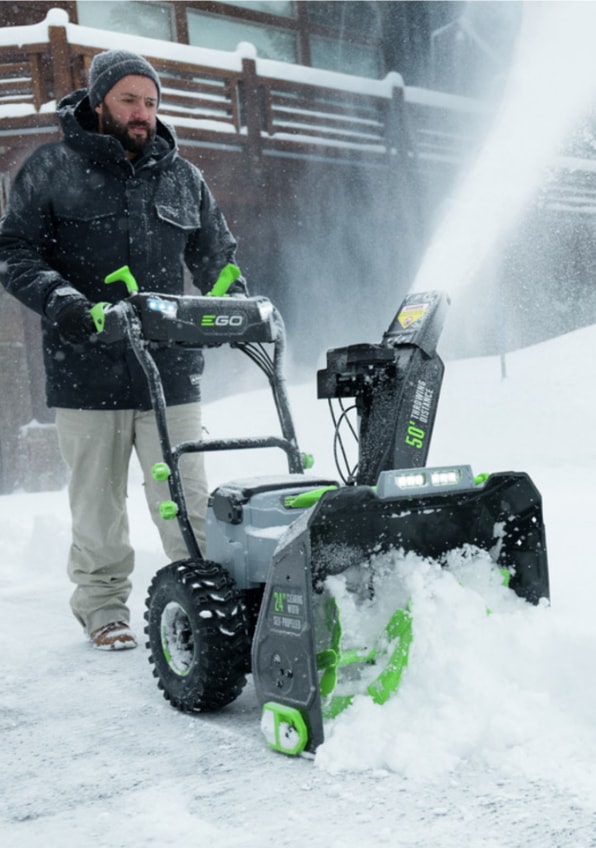 A person operates a snowblower, clearing snow from a driveway or pathway in a snowy environment.