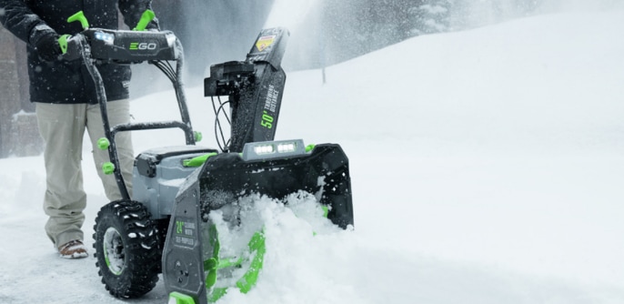 A person operates a snowblower, clearing snow from a driveway or pathway in a snowy environment.