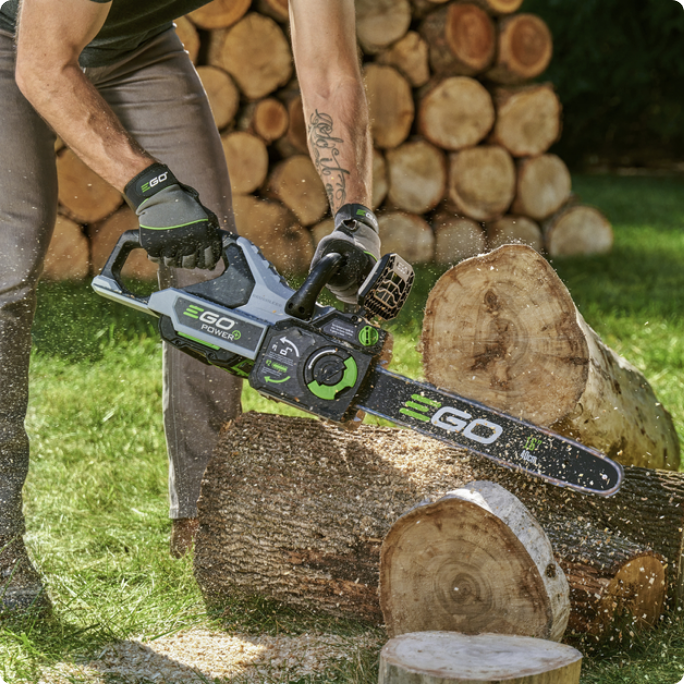 A person uses an EGO chainsaw to cut a log, with a stack of cut firewood in the background.