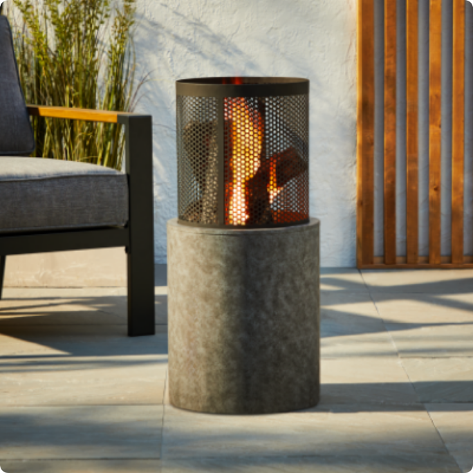 A small outdoor fire pit with a cylindrical base and mesh cover, placed on a patio near a chair.