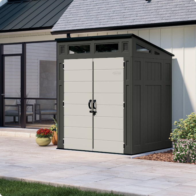 A small, modern outdoor storage shed with double doors and a slanted roof, situated on a patio beside a house.
