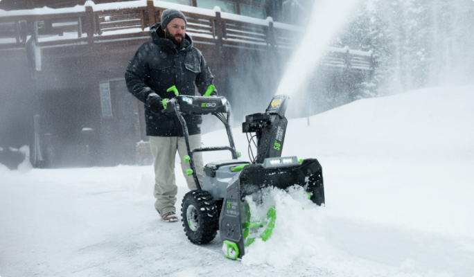 A man pushing a grey and green snowblower in a snowy driveway.