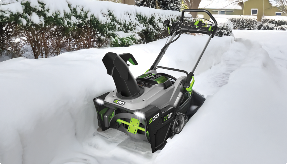 A cordless grey and green snowblower clearing a walkway filled with snow.