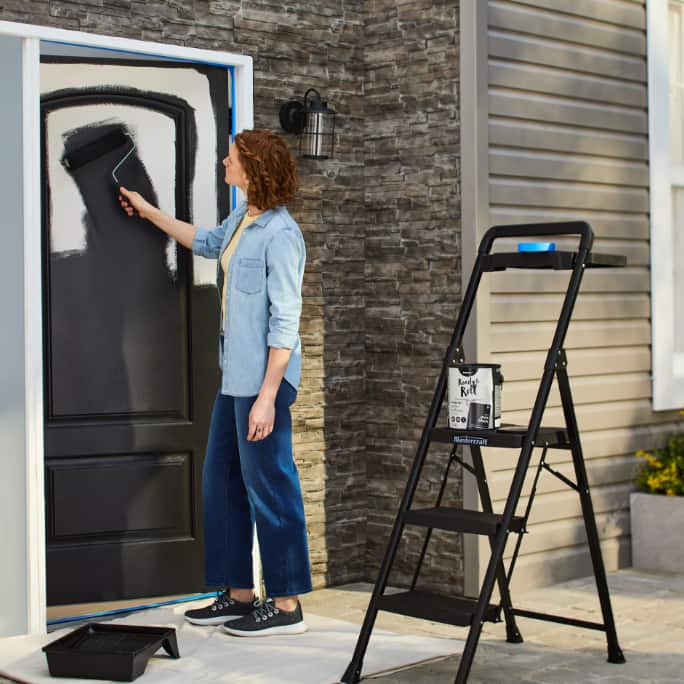 A woman standing beside a step ladder painting a black front door.