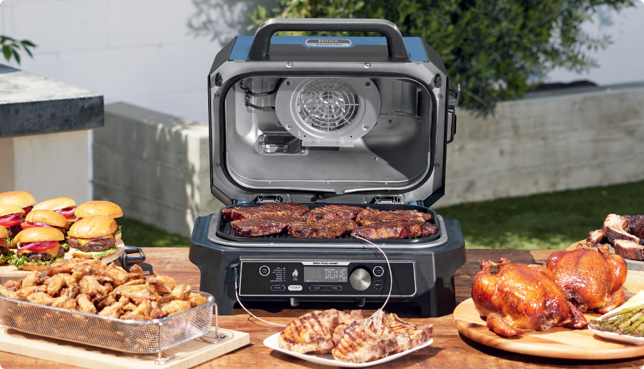A tabletop grill cooking steaks on a table, next to various prepared foods.