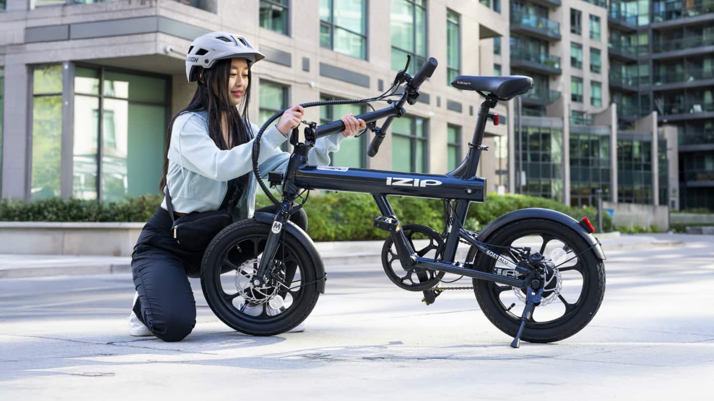 Woman with a helmet adjusting her iZip eBike in an urban setting. Text reads “iZip eBikes & eScooters" and includes a "Shop Now" button.