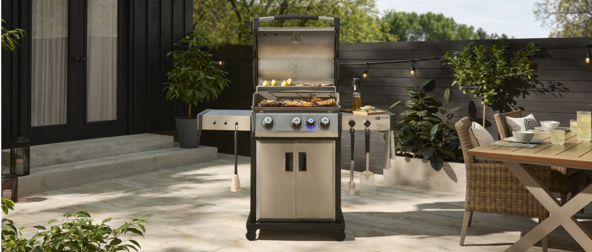 Un barbecue au gaz en acier inoxydable avec le couvercle ouvert sur lequel cuisent des aliments, sur une spacieuse terrasse pavée de pierre. La terrasse comprend des meubles d’extérieur et est entouré de verdure et de plantes en pot, avec une clôture en bois foncé en arrière-plan.