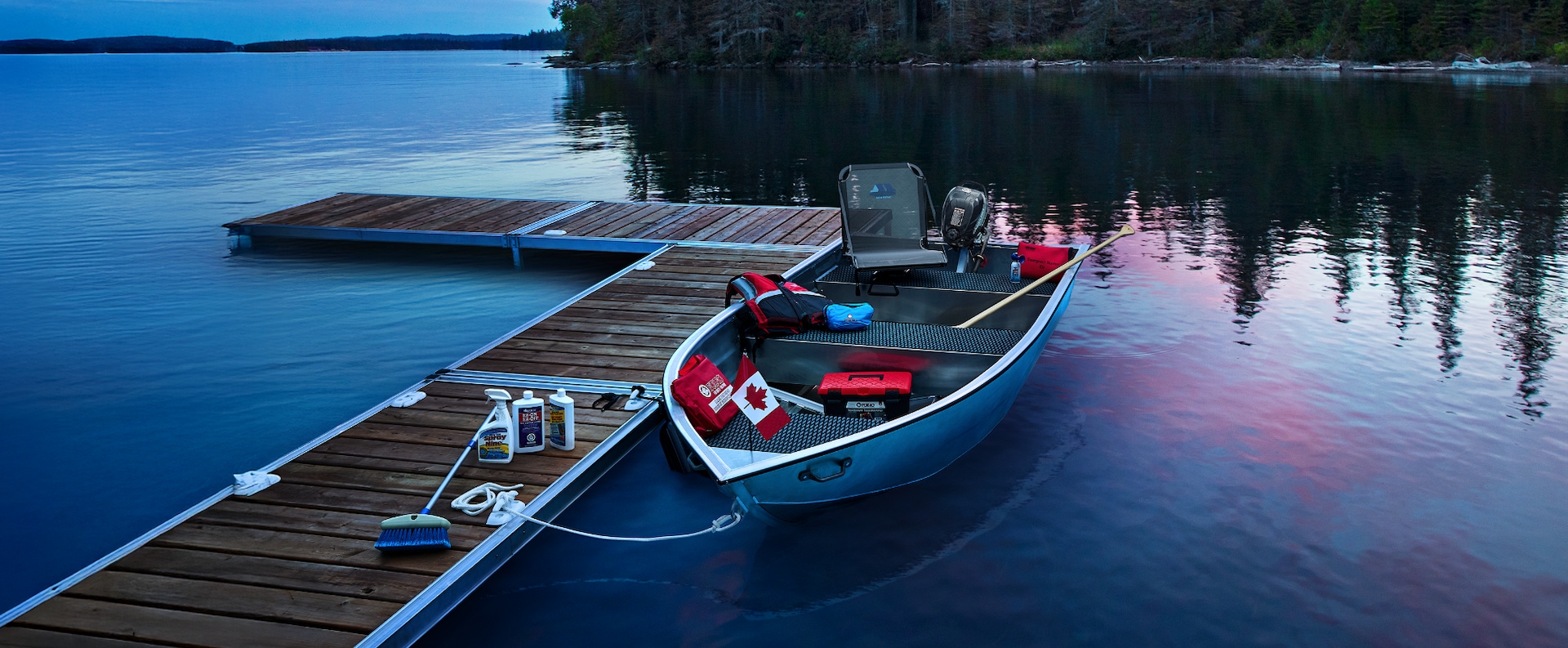 A small fishing boat is tied to a wooden dock on a calm lake at dusk, with trees and distant shoreline visible in the background.