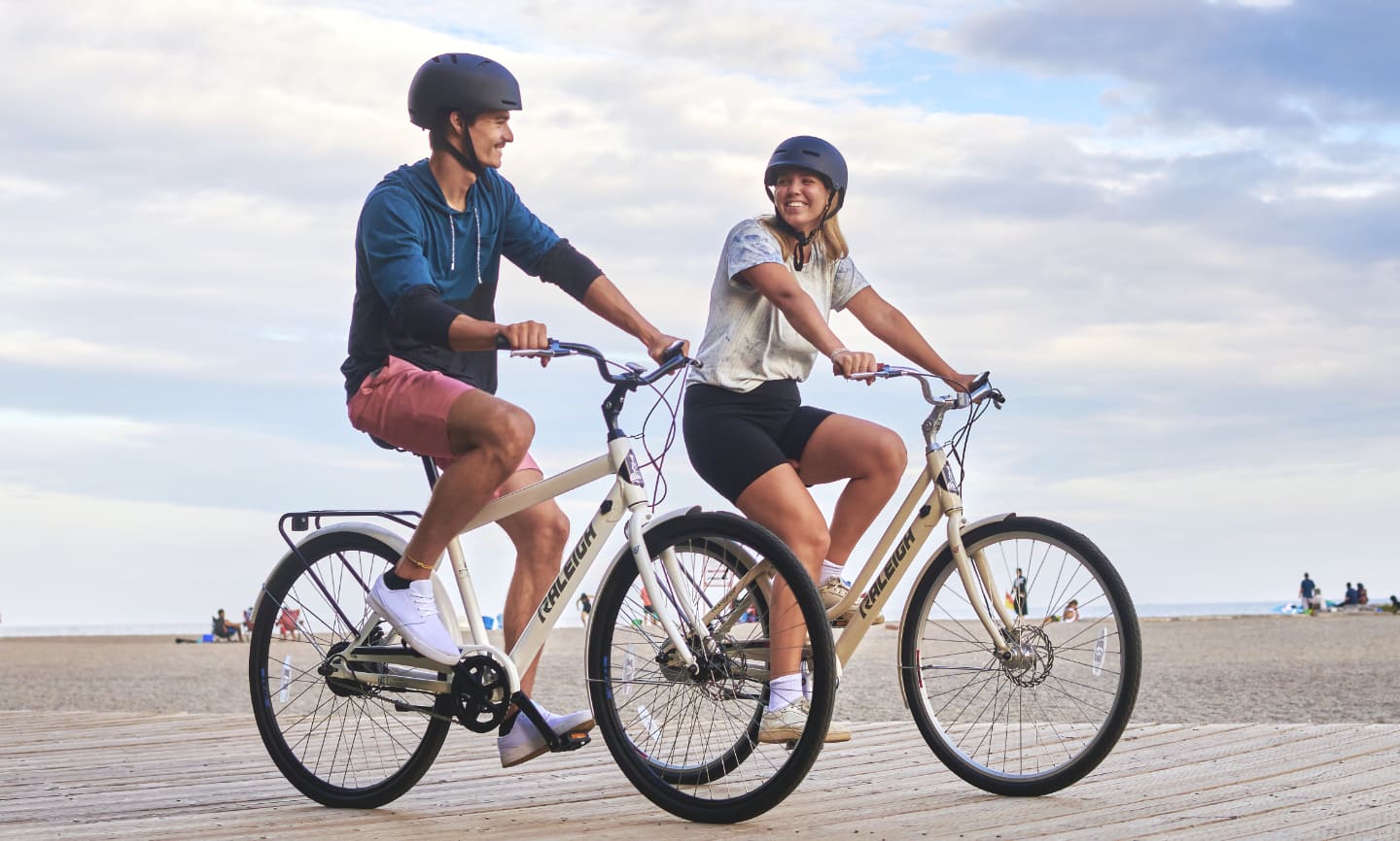 Two adults riding Raleigh bikes on a beach boardwalk. 