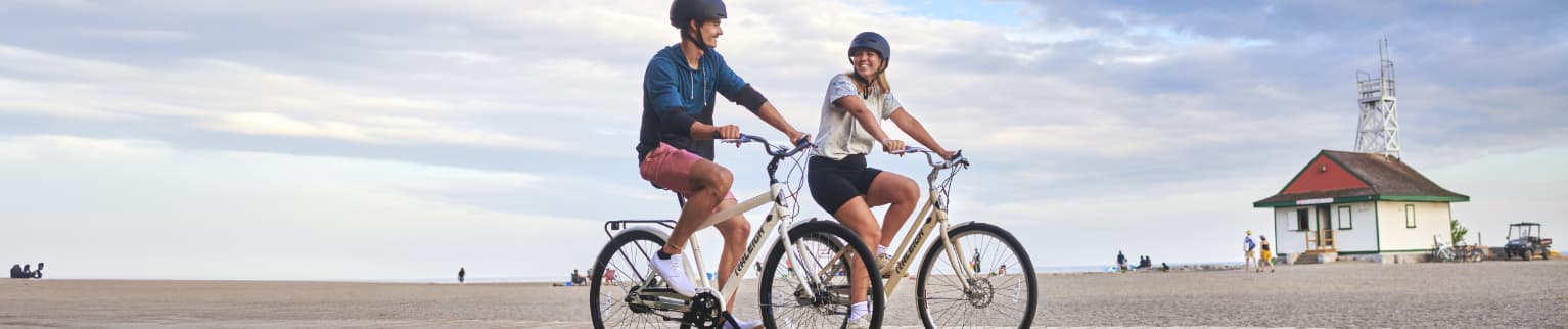 Two adults riding Raleigh bikes on a beach boardwalk. 