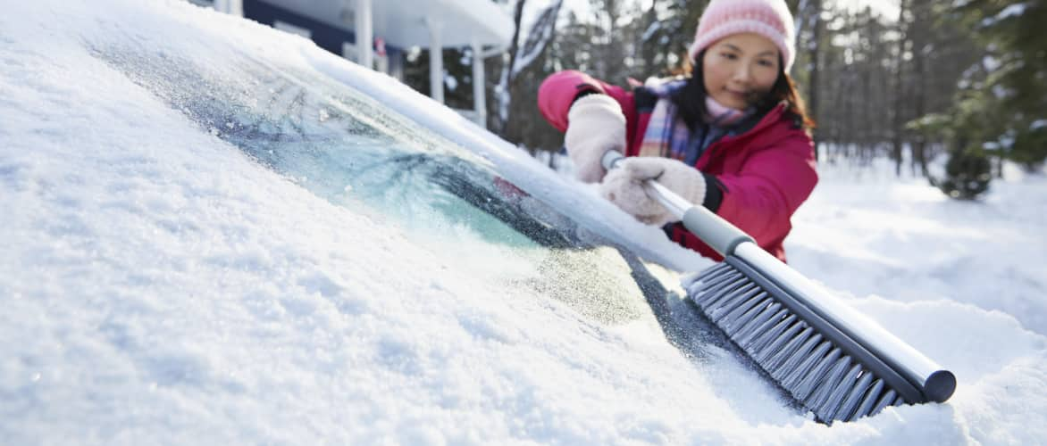 A woman clears snow from a car windshield with a snow brush.